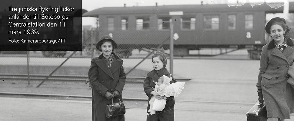 svartvitt foto av tre flickor i kappor a la 1930-tal vid järnvågsspår. Den i mitten bär overall smat håller en dock i famnen.